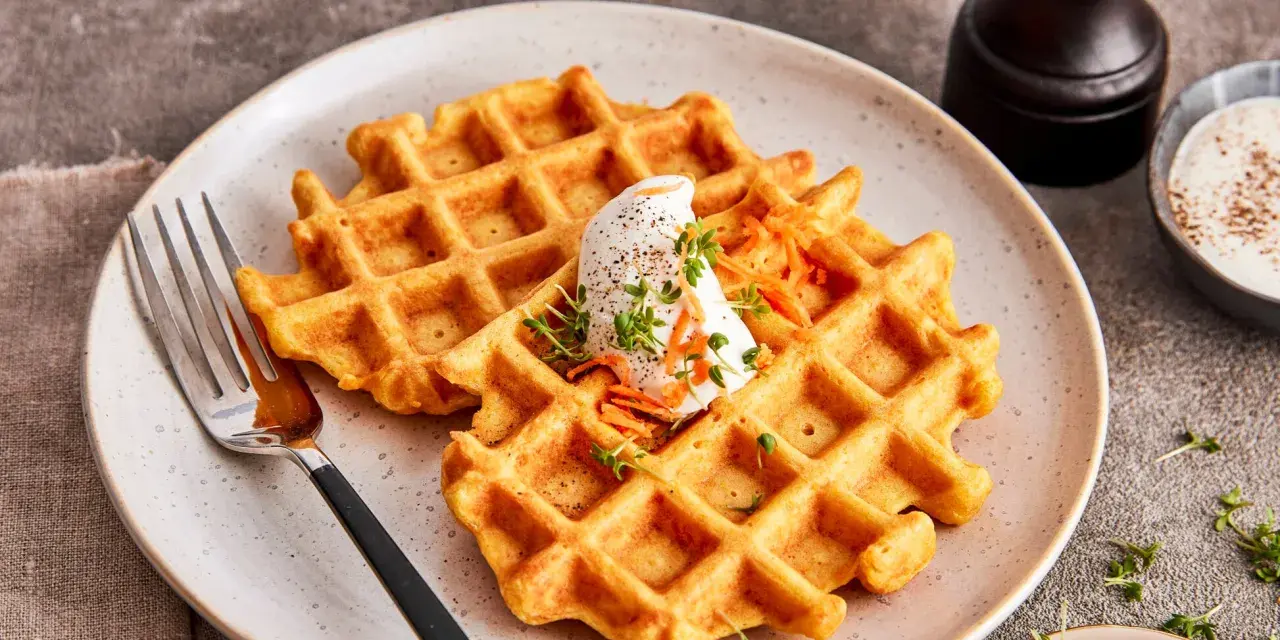 Zwei goldbraune Karotten Waffeln mit einem Klecks Sahne und geraspelten Karotten, garniert mit Kresse.