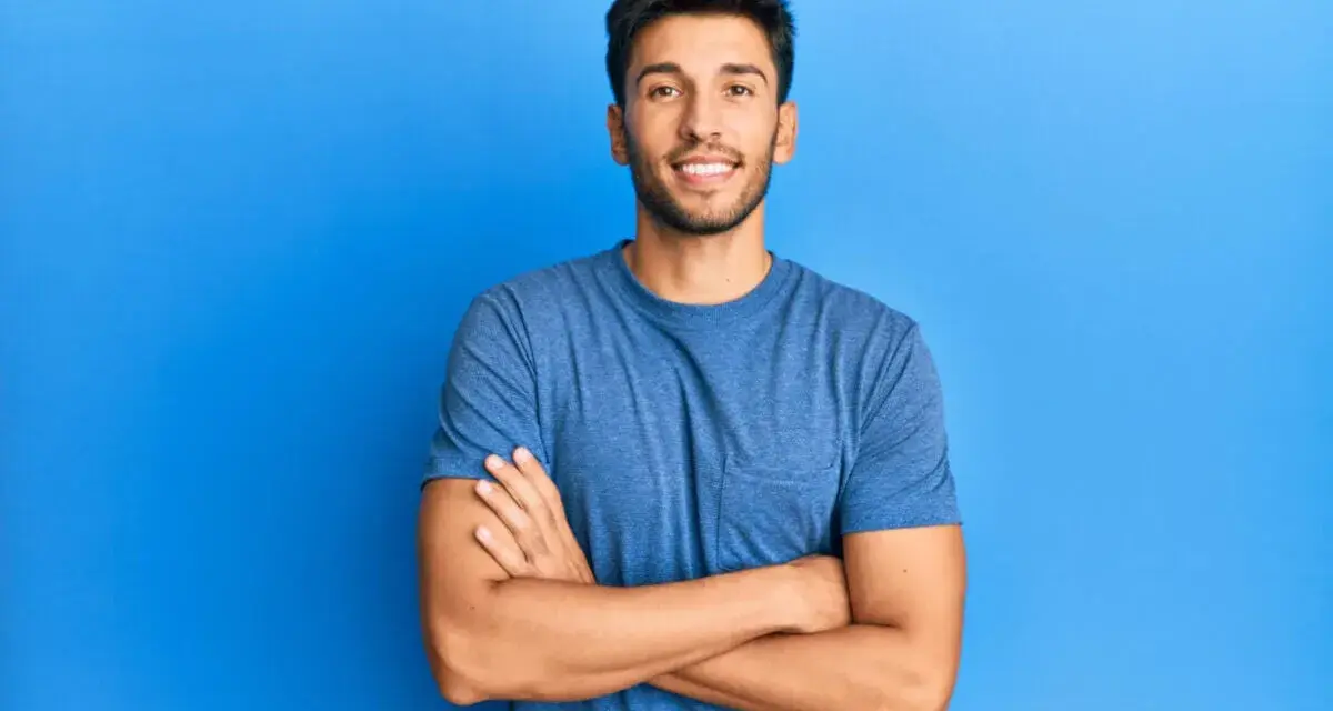Hombre sonriente con estilos barba poco poblada, brazos cruzados, sobre fondo azul.