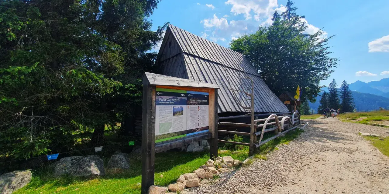 Drewniana chata na Rusinowej Polanie, obok tablica informacyjna o wypasie owiec. W tle góry i błękitne niebo.