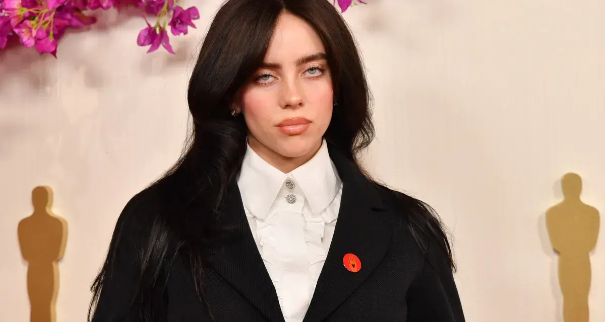 Billie Eilish z charakterystycznymi sanpaku eyes, w czarnym żakiecie i białej koszuli, na tle ozdobnych kwiatów i złotych statuetek.