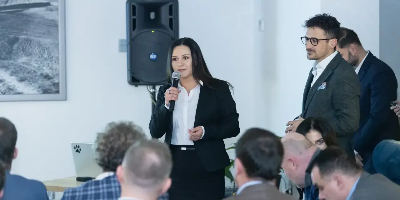 Kobieta w garniturze przemawia do grupy ludzi, obok mężczyzna w okularach.
