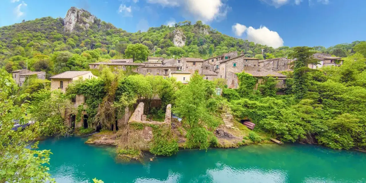 Un borgo incantevole in Umbria, con case in pietra ricoperte di vegetazione che si affacciano su un fiume turchese. Un luogo perfetto per i cammini.