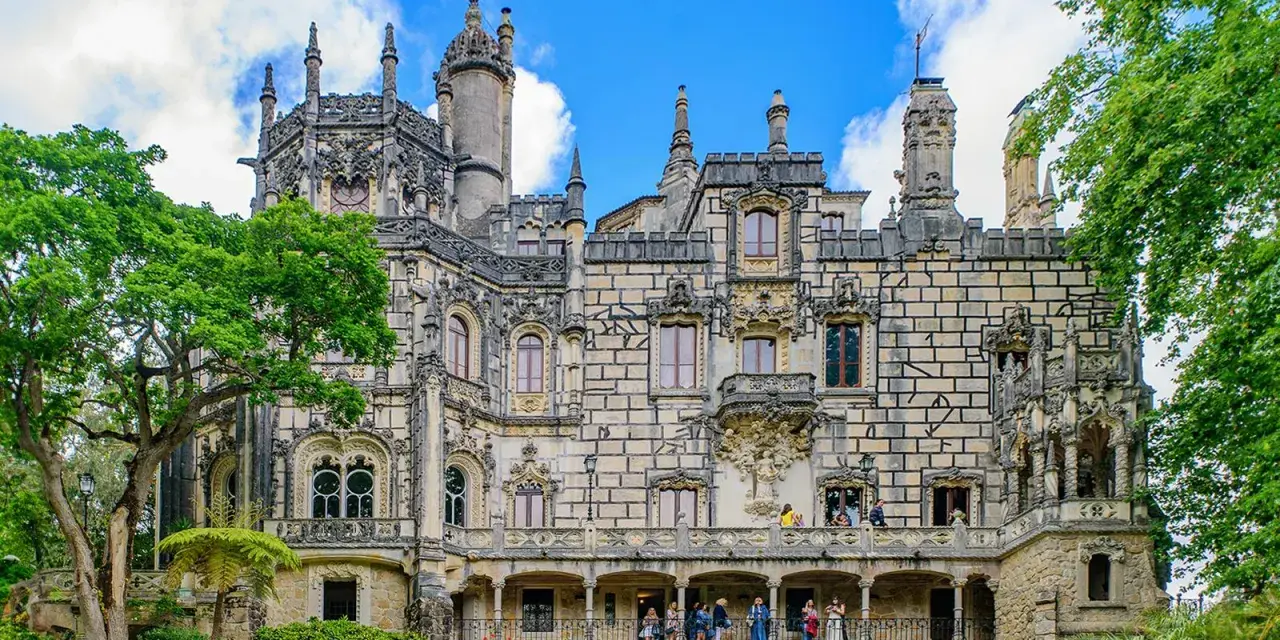 The Quinta da Regaleira estate in Sintra, Portugal, features a stunning palace with intricate stonework and ornate balconies, surrounded by lush greenery under a blue sky.