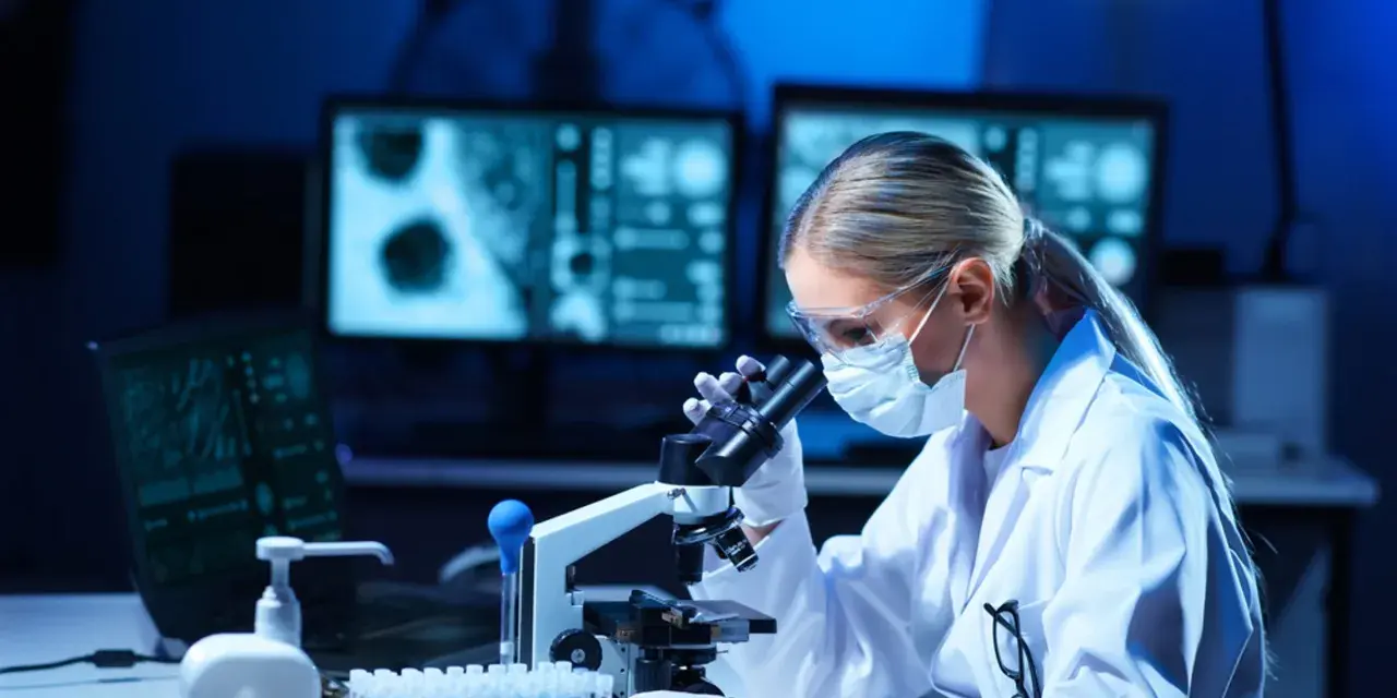 Kobieta w masce i okularach ochronnych pracuje przy mikroskopie w laboratorium.