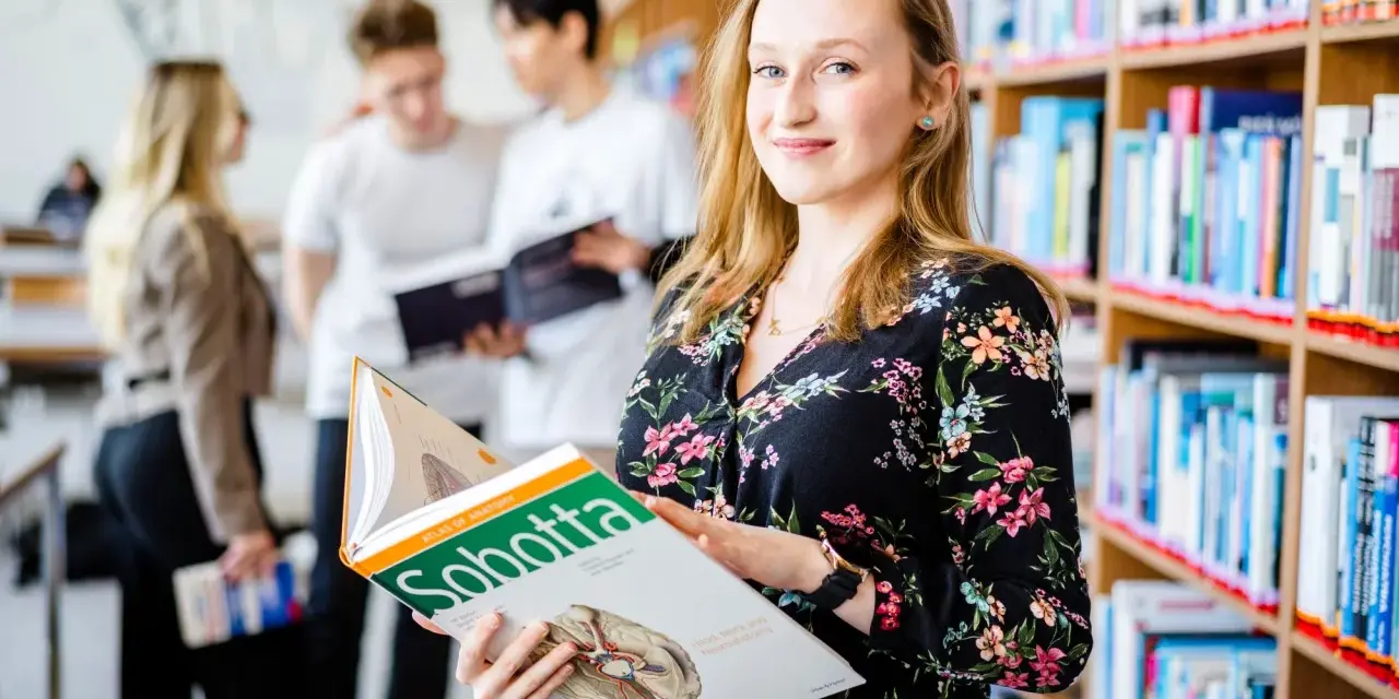 Studentka na Uniwersytecie Wrocławskim, kierunek prawo, przegląda atlas anatomii. W tle inni studenci i regały z książkami.