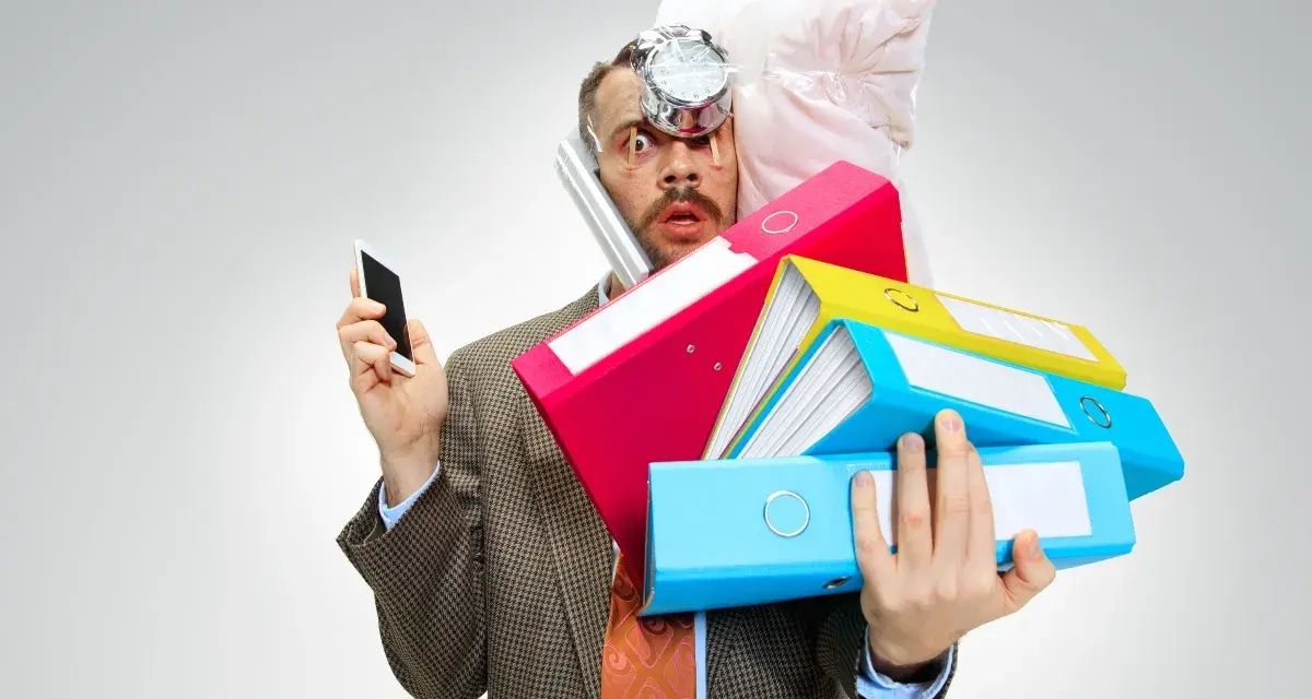 Uomo stressato con pile di cartelle, telefono e cuscino in testa. Lo stress aumenta il cortisolo, che fa ingrassare.