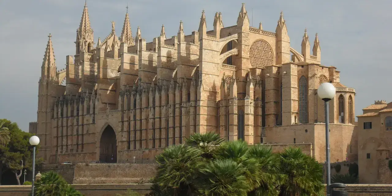 La cathédrale de Palma, majestueuse, se dresse sous un ciel d'hiver. Des palmiers verdoyants bordent le chemin, offrant un contraste saisissant avec la pierre dorée du monument.