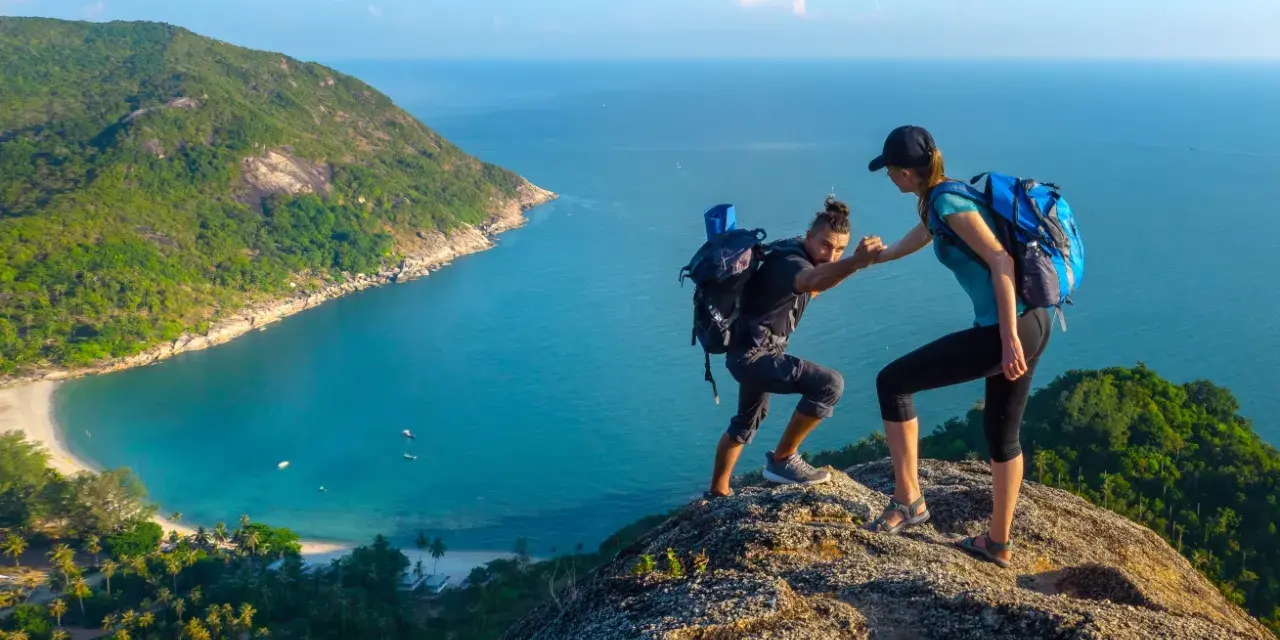 Due escursionisti si aiutano a salire su una scogliera con vista su una baia tropicale. Questo è il trekking cos'è: avventura e collaborazione.