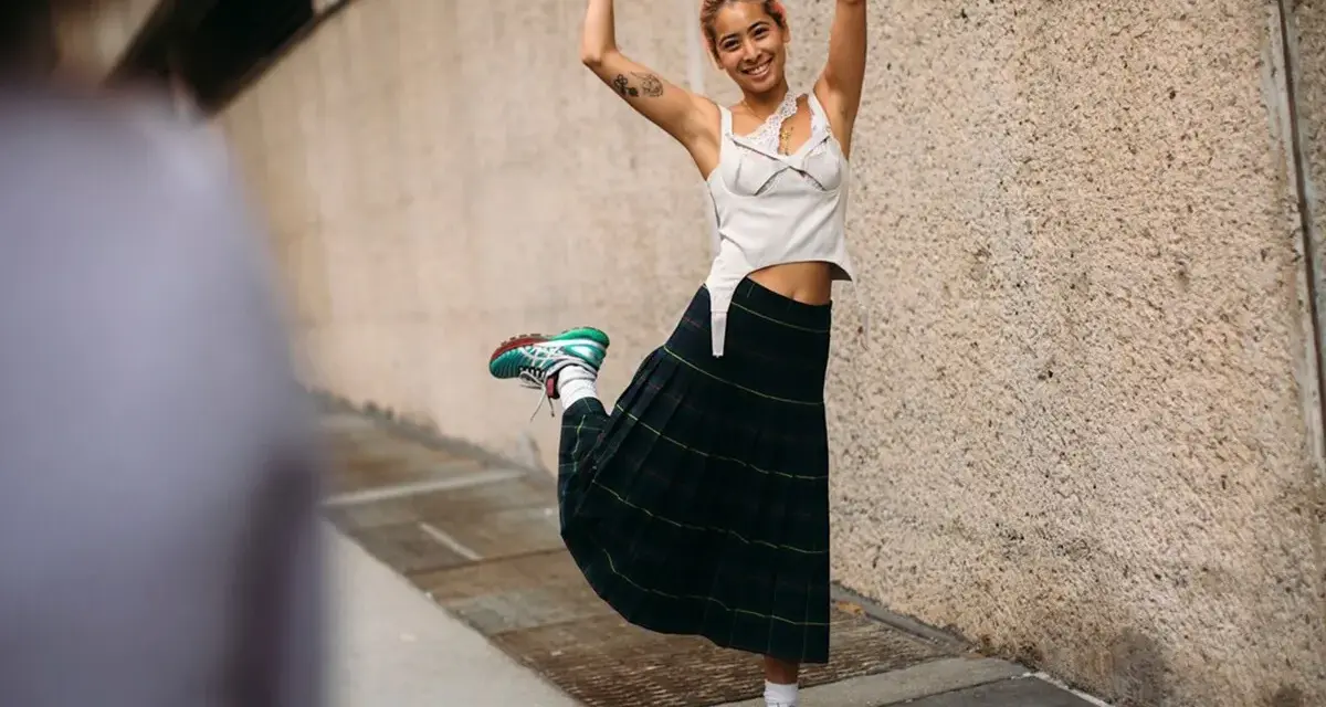 Joven sonriente con falda escocesa y top blanco, luciendo unos modernos looks con zapatillas deportivas.