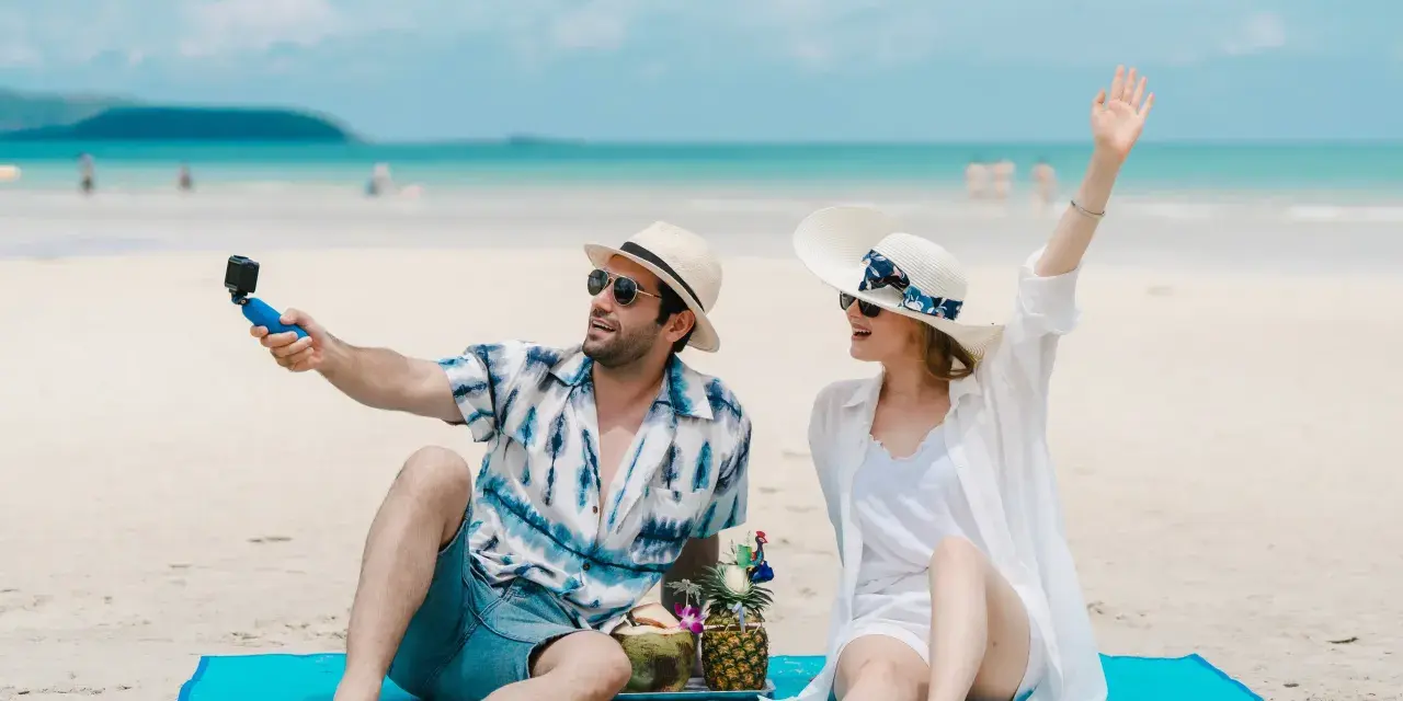 Para na plaży nagrywa filmik kamerką GoPro, a jego partnerka macha do obiektywu. Idealne wakacje pod gruszą!
