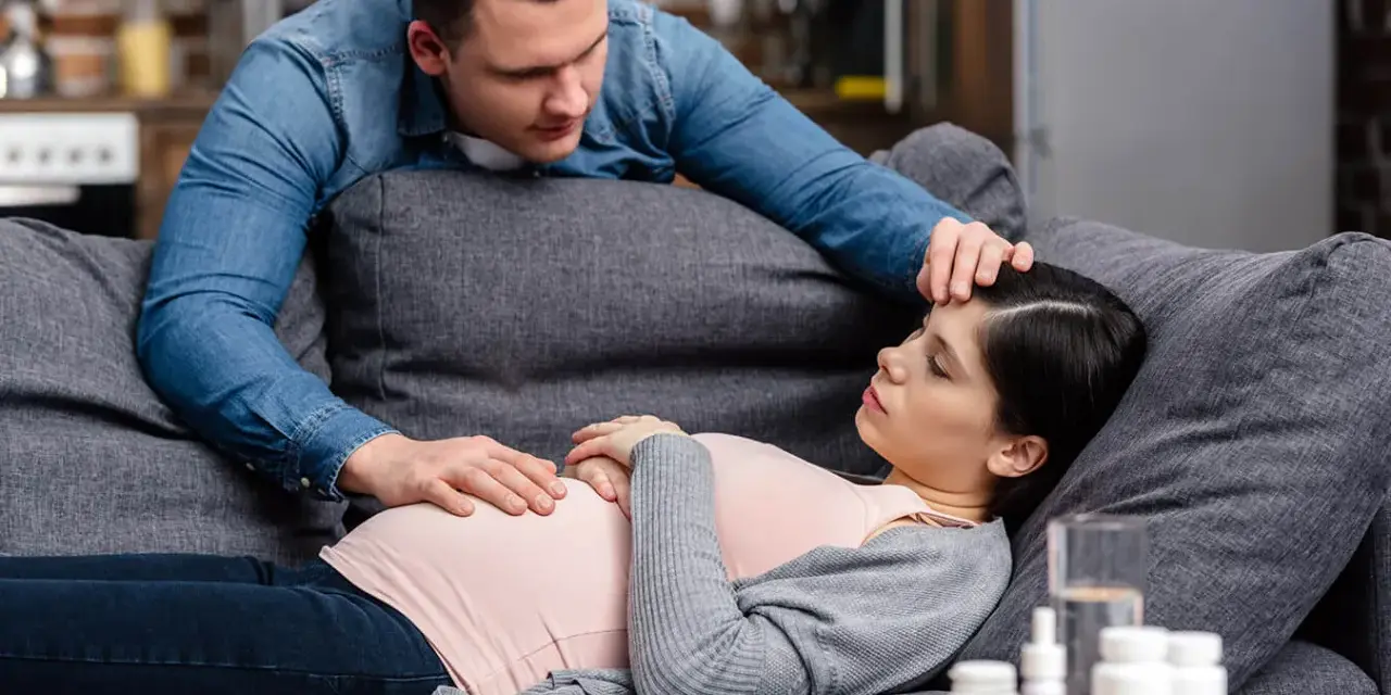Zmartwiony partner troskliwie dotyka czoła ciężarnej kobiety, która leży na kanapie. Na stoliku leki, sugerujące problemy zdrowotne, być może angina w ciąży.