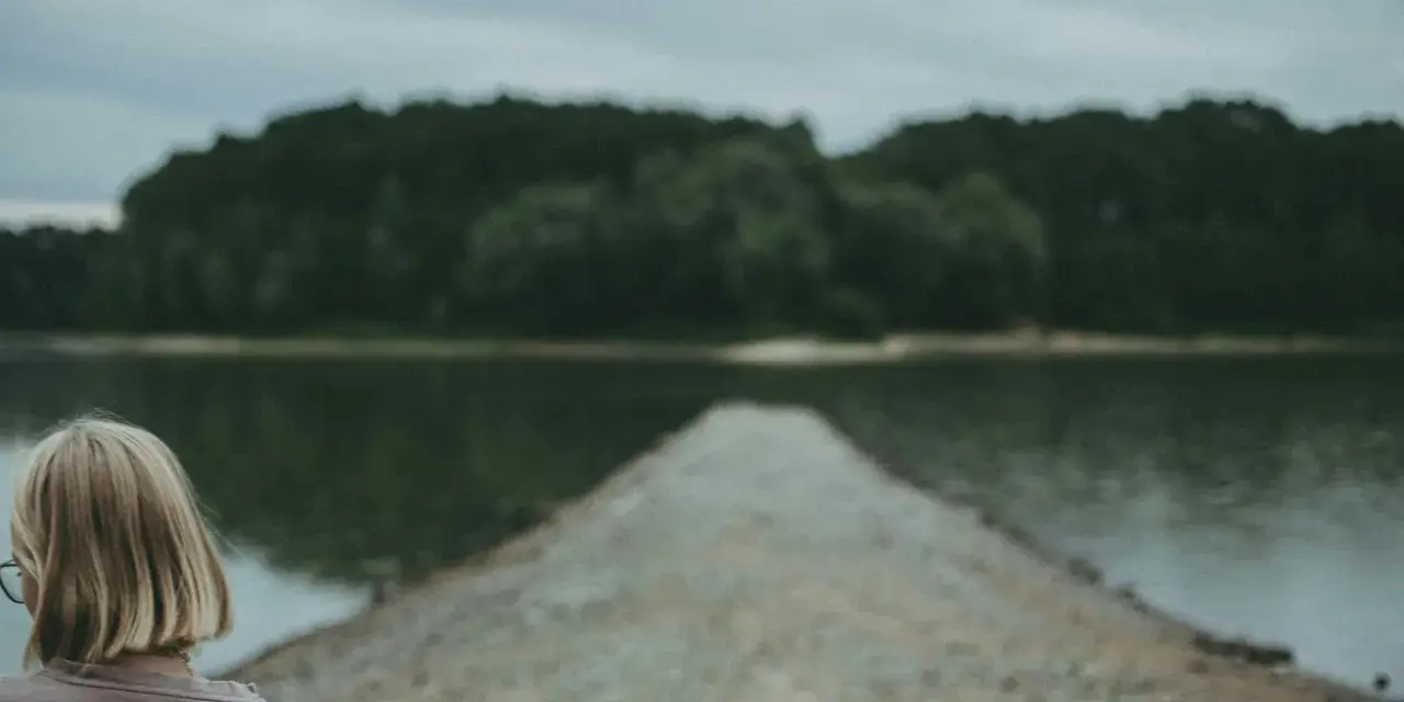 Dziewczyna z blond włosami patrzy na spokojną taflę jeziora, czując uczucie pustki.