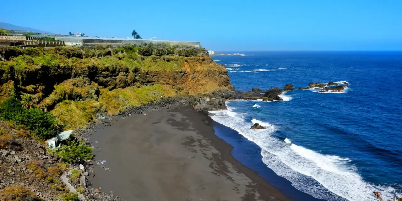 Spiaggia di sabbia nera e scogliere verdi sulle isole delle Canarie, con il blu intenso dell'oceano Atlantico.