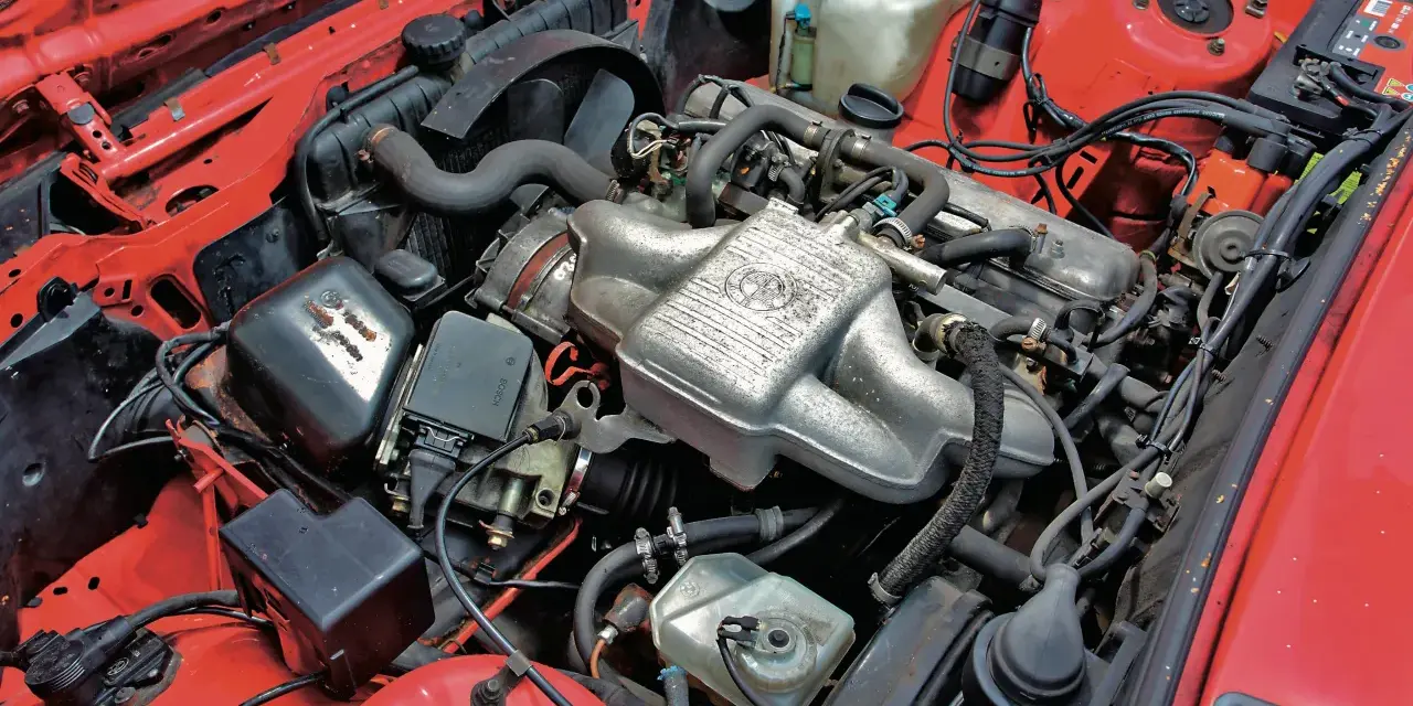 Silniki BMW E30 w czerwonej komorze silnika. Widoczny kolektor ssący z logo BMW, chłodnica, akumulator i inne elementy układu napędowego.