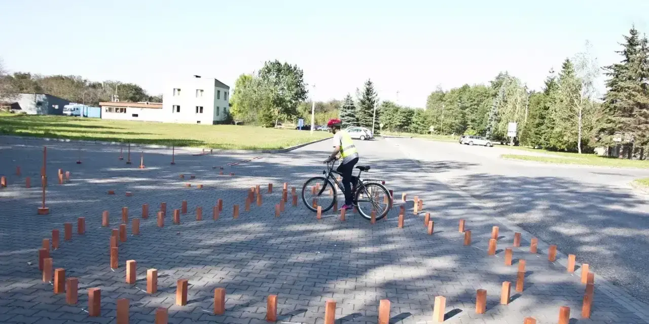 Młody rowerzysta w kamizelce odblaskowej i kasku pokonuje tor przeszkód z palików, przygotowując się do egzaminu na kartę rowerową.