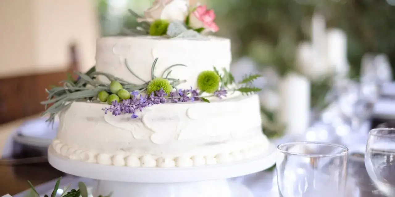 Dwupoziomowy tort weselny ozdobiony kwiatami, ziołami i oliwkami. Delikatne kremowe wykończenie i naturalne dekoracje podkreślają wyjątkowe smaki tortów weselnych.