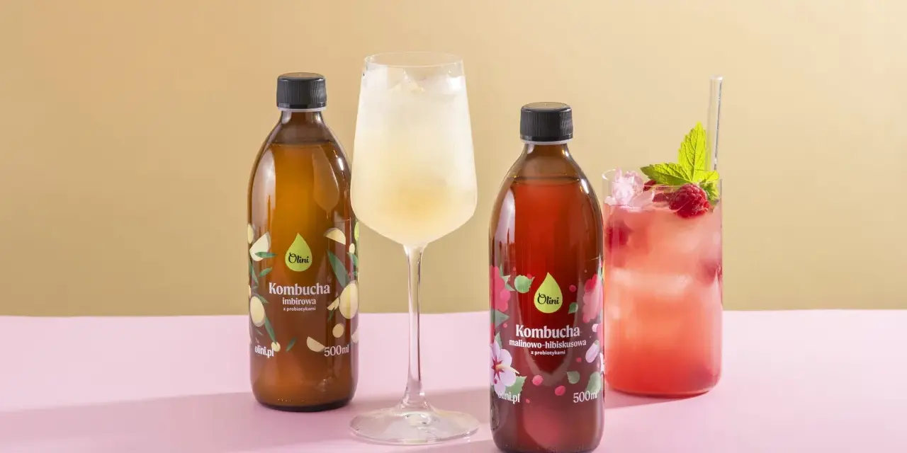 Kombucha co to? Dwie butelki kombuchy Olini: imbirowej i malinowo-hibiskusowej, obok orzeźwiających napojów z lodem.