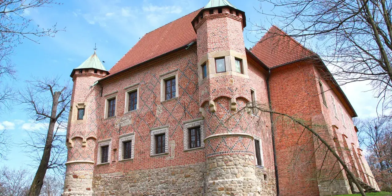 Zamek w Dębnie, ceglana twierdza z kamienną podstawą i zielonymi wieżyczkami, otoczony drzewami pod błękitnym niebem.