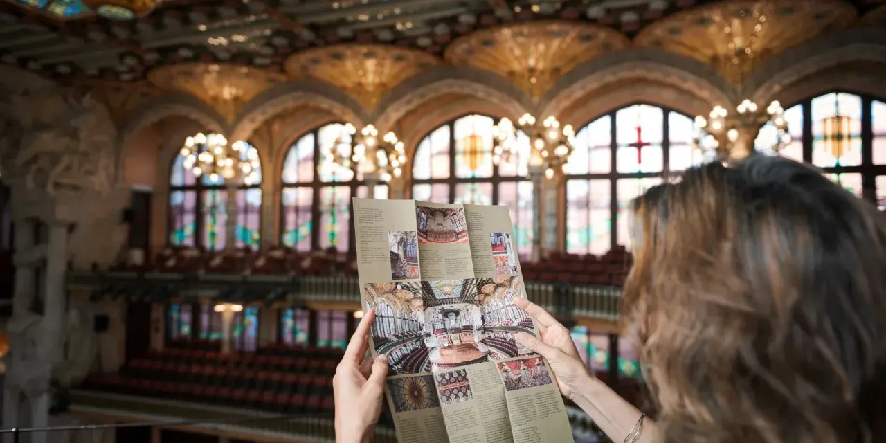 Kobieta studiuje broszurę z widokiem na wspaniałe wnętrze Pałacu Muzyki Katalońskiej, z witrażowym sufitem i rzędami siedzeń.