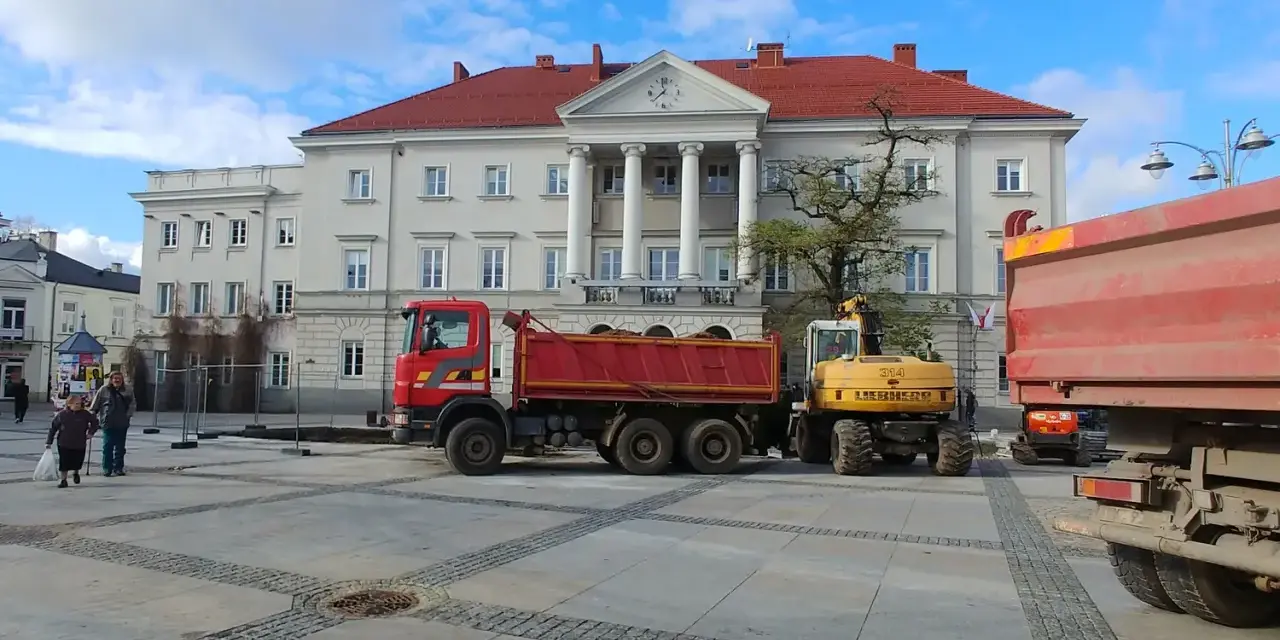 Czerwona wywrotka i żółta koparka na placu przed budynkiem z kolumnami.