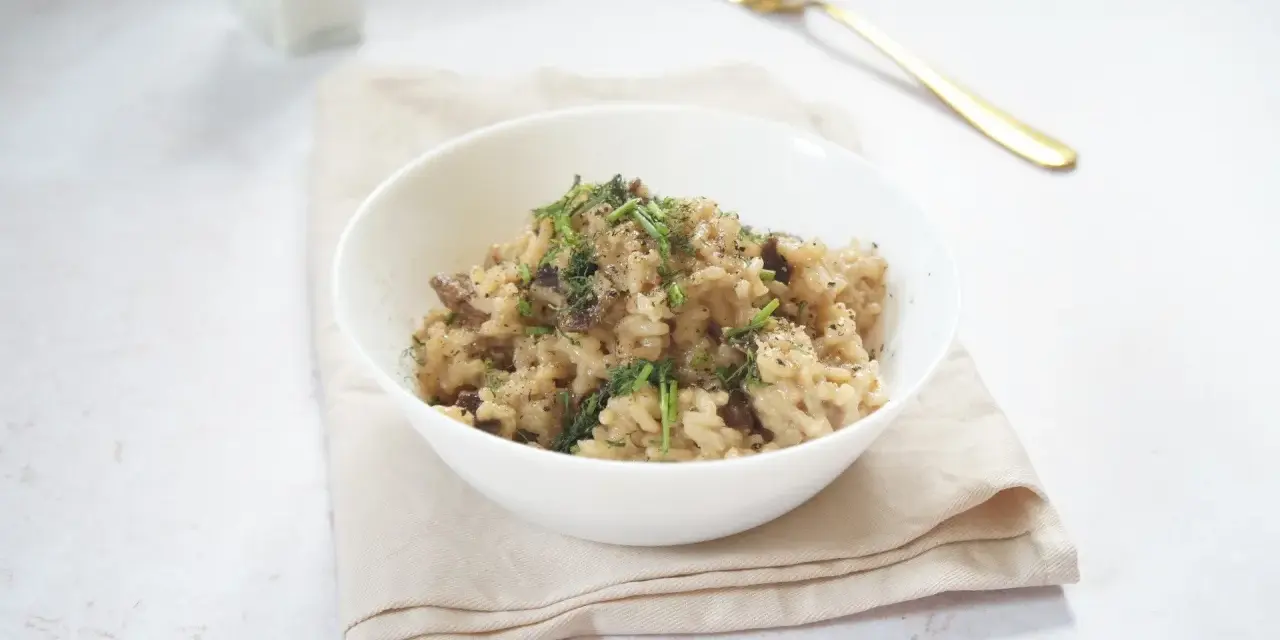 Miseczka kremowego risotto z grzybami, posypana ziołami i pieprzem, obok złoty widelec.