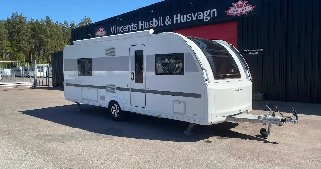 En vit Adria Alpina 613 UT husvagn står parkerad framför en byggnad med texten "Vincents Husbil & Husvagn".