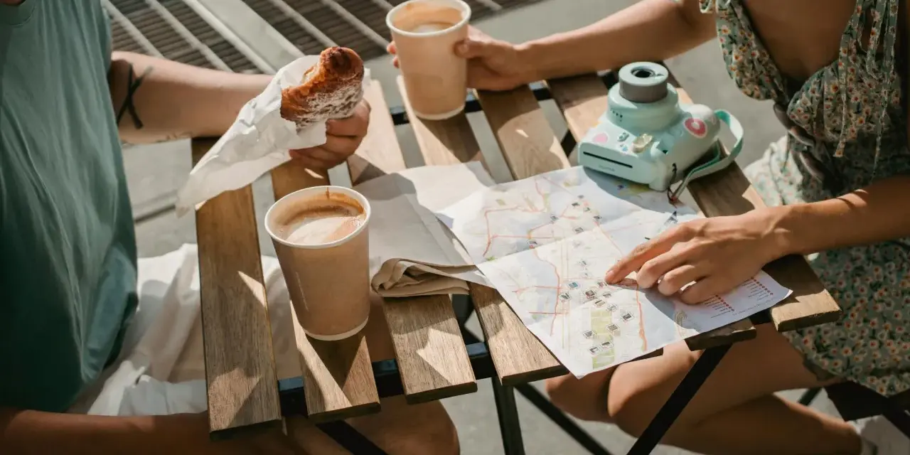 Para planuje podróż, studiując mapę i jedząc croissanty. Na stole kawa i aparat.
