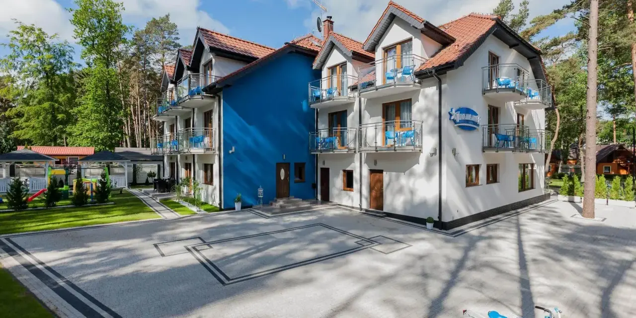 Idealne wczasy nad morzem z wyżywieniem. Biały budynek z niebieskimi akcentami, tarasy z widokiem na las, plac zabaw i rowery czekają na gości.