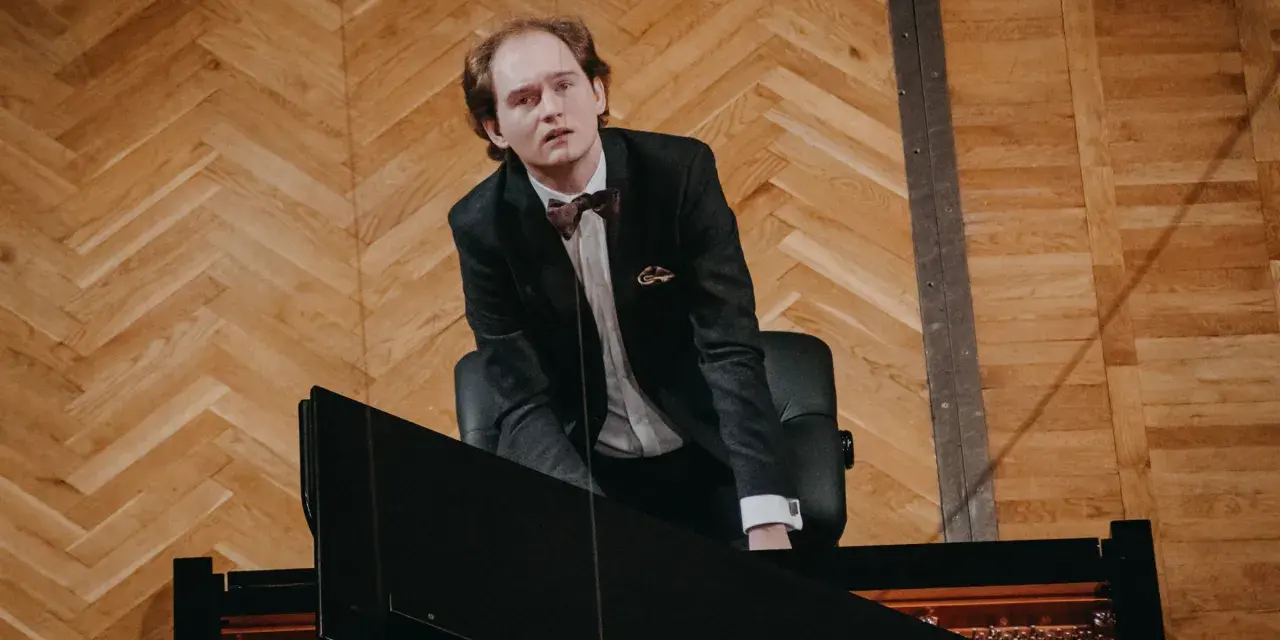 Piotr Ryszard Pawlak w eleganckim garniturze pochyla się nad fortepianem, gotowy do wykonania utworu.