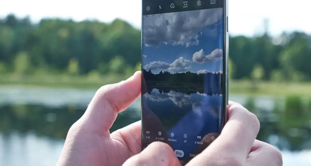 Dłonie trzymają smartfon Samsung S10, na ekranie którego widać odbicie nieba i lasu w spokojnej wodzie.