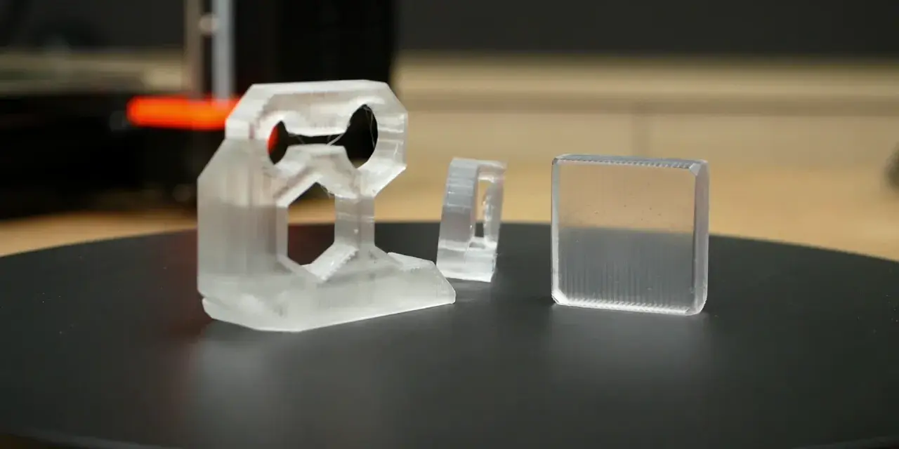 Three clear 3D printed objects, one complex bracket and two simpler shapes, sit on a black surface.