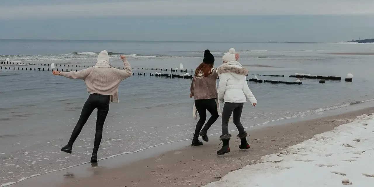 Trzy dziewczyny cieszą się zimowym dniem, gdzie zimą nad morze. Spacerują po plaży, na której leży śnieg, a fale delikatnie rozbijają się o brzeg.