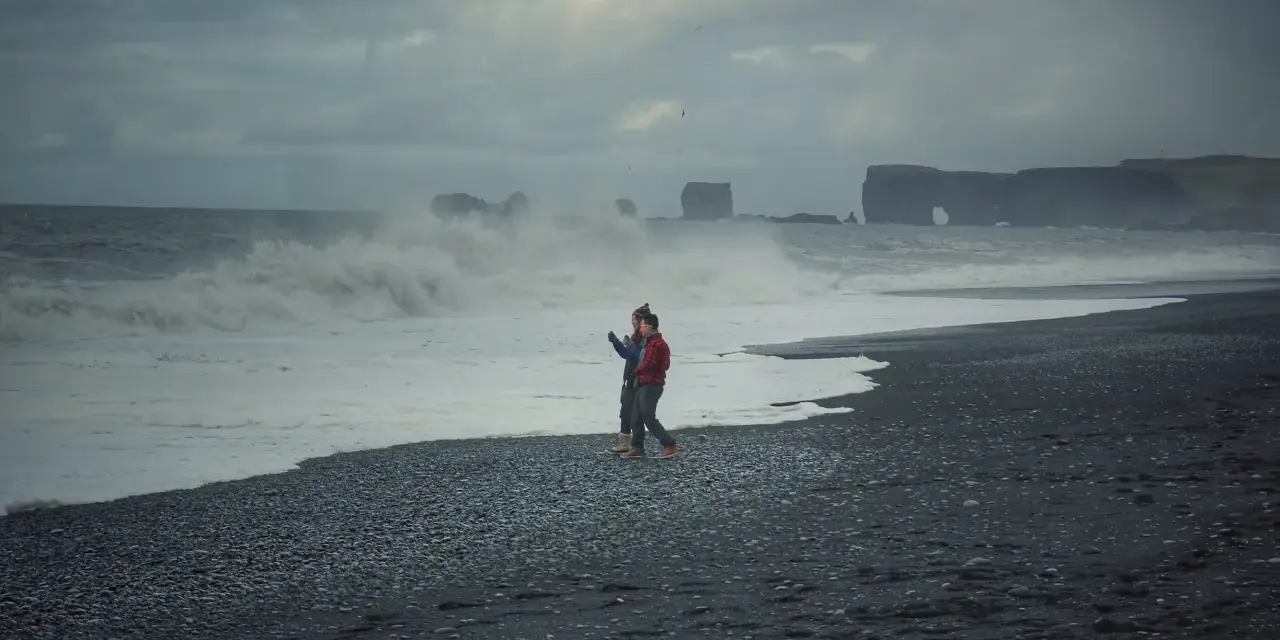 Para spaceruje po czarnej plaży Islandii, podczas gdy fale rozbijają się o brzeg.