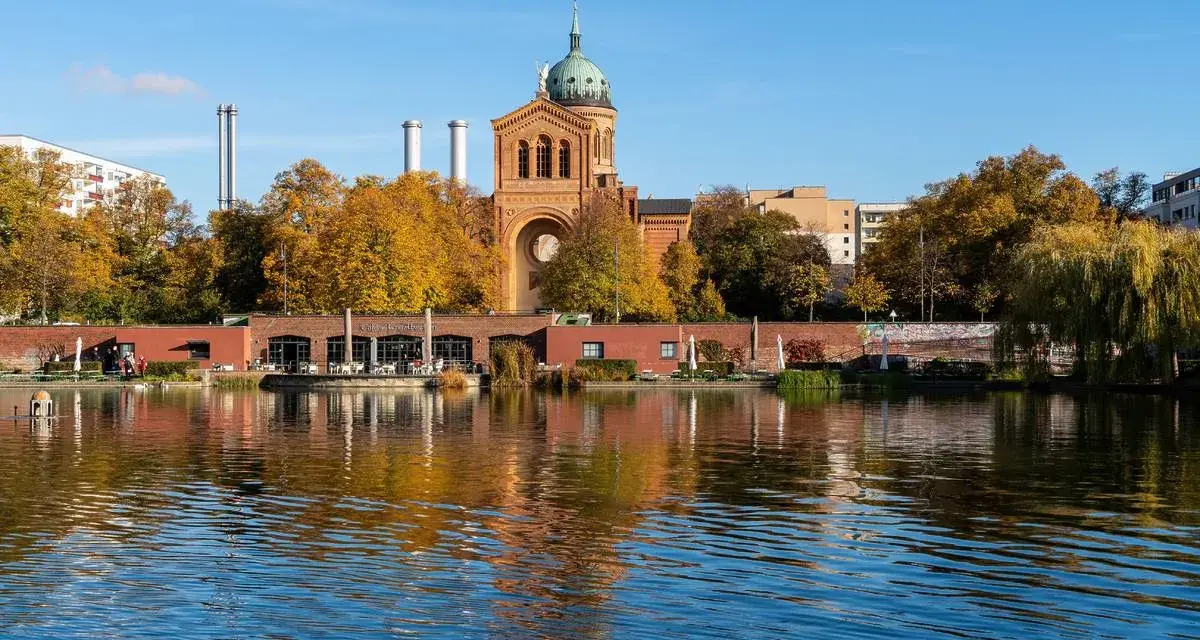 Herbstliche Bäume spiegeln sich im Wasser des Engelbeckens Berlin. Ein Café mit Außenbereich und eine Kirche mit Kuppel sind zu sehen.