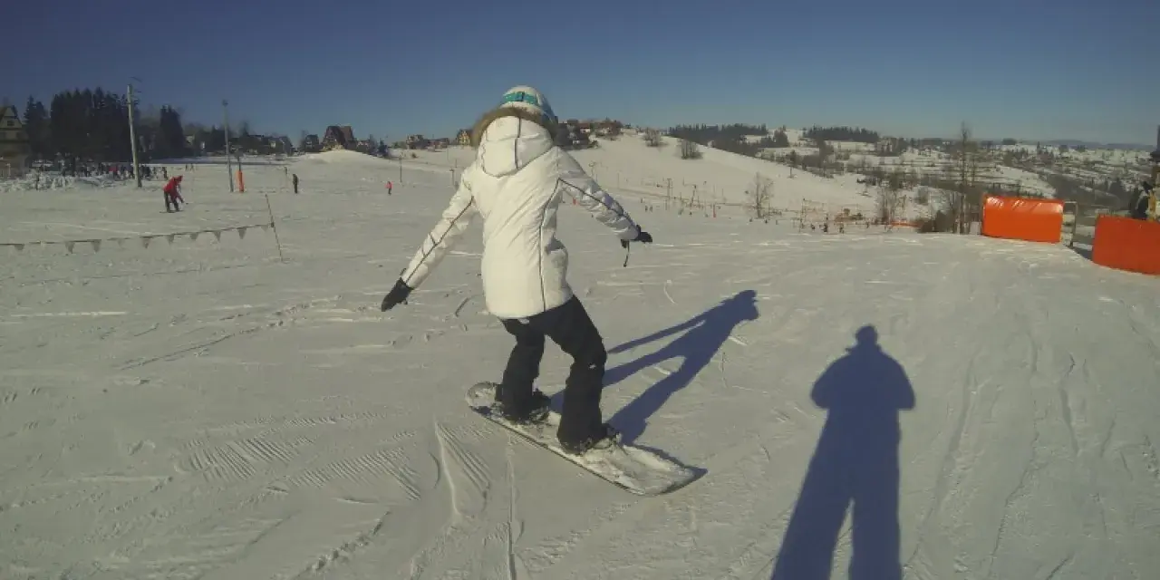 Osoba na snowboardzie na stoku narciarskim, ucząca się w szkole narciarskiej Budzyńskich. Słoneczny dzień, czyste niebo.