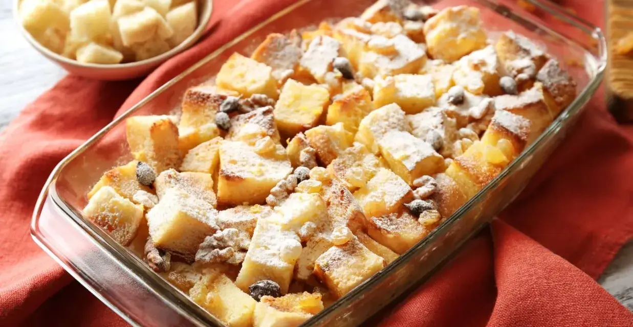 Pudding chlebowy w naczyniu, posypany cukrem pudrem i rodzynkami, obok miseczka z pokrojonym chlebem.