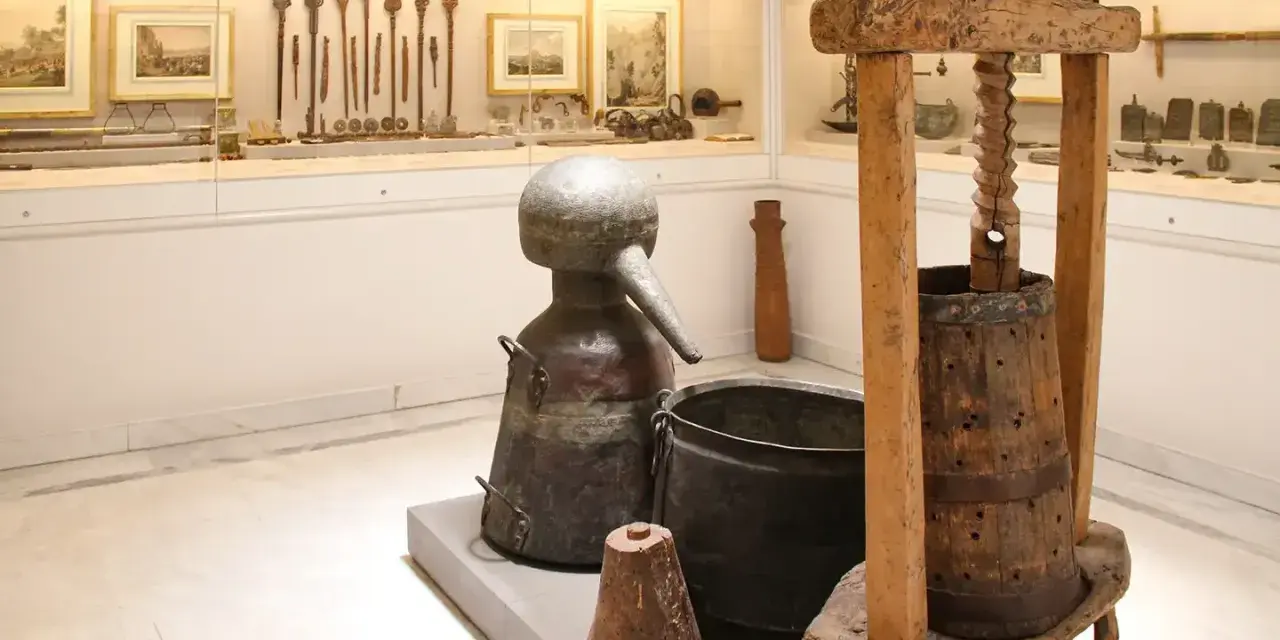 W muzeum Ateny: zabytkowy alembik, kadź i prasa do winogron. W tle ekspozycja narzędzi i obrazów.