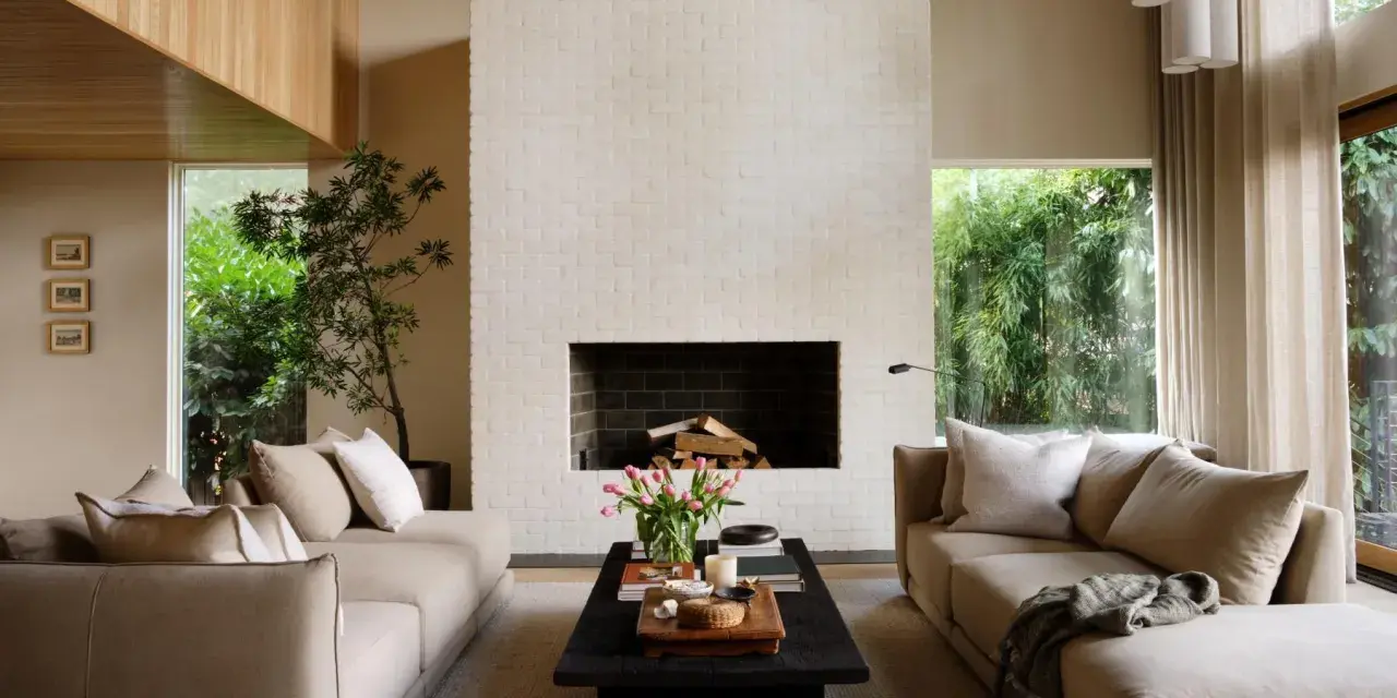 Salón de estilo japandi con sofás beige, chimenea de ladrillo blanco y mucha luz natural.