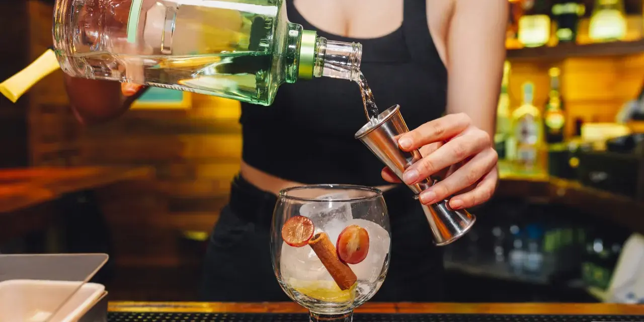 Barman nalewa alkohol z butelki do miarki nad kieliszkiem z lodem i owocami.