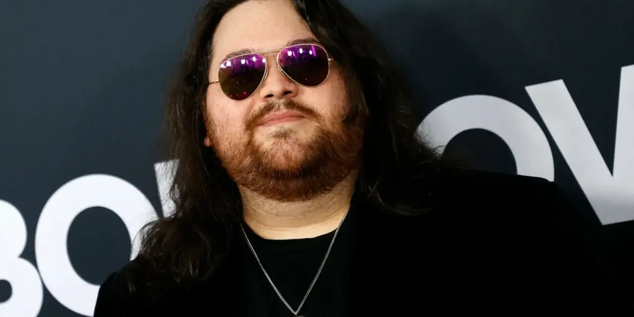 Wolfgang Van Halen w okularach przeciwsłonecznych i z naszyjnikiem w kształcie serca, na tle logo HBO.