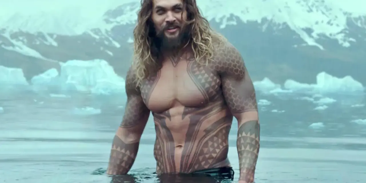 Jason Momoa w wodzie, z tatuażami, jak w filmach i serialach. W tle góry i lód.