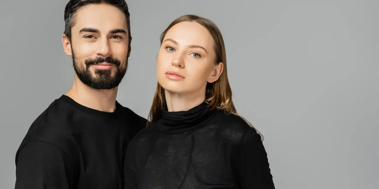 Para, która wygląda jakby mogła nosić ubrania unisex. Mężczyzna z brodą i kobieta z długimi włosami, oboje w czarnych, prostych ubraniach.