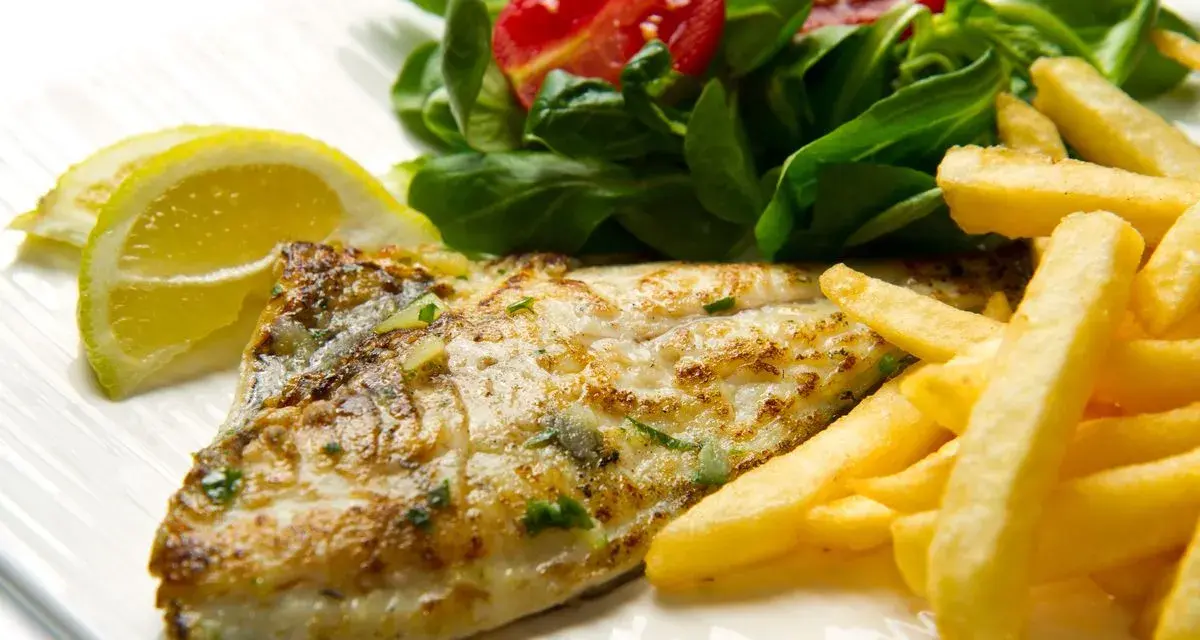 A cooked fish fillet with french fries, salad, and lemon slices on a white plate.