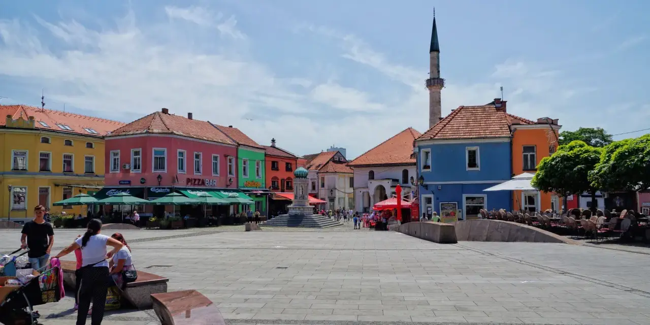 Kolorowy plac w **Tuzla, Bośnia**, z kawiarniami, fontanną i minaretem w tle.