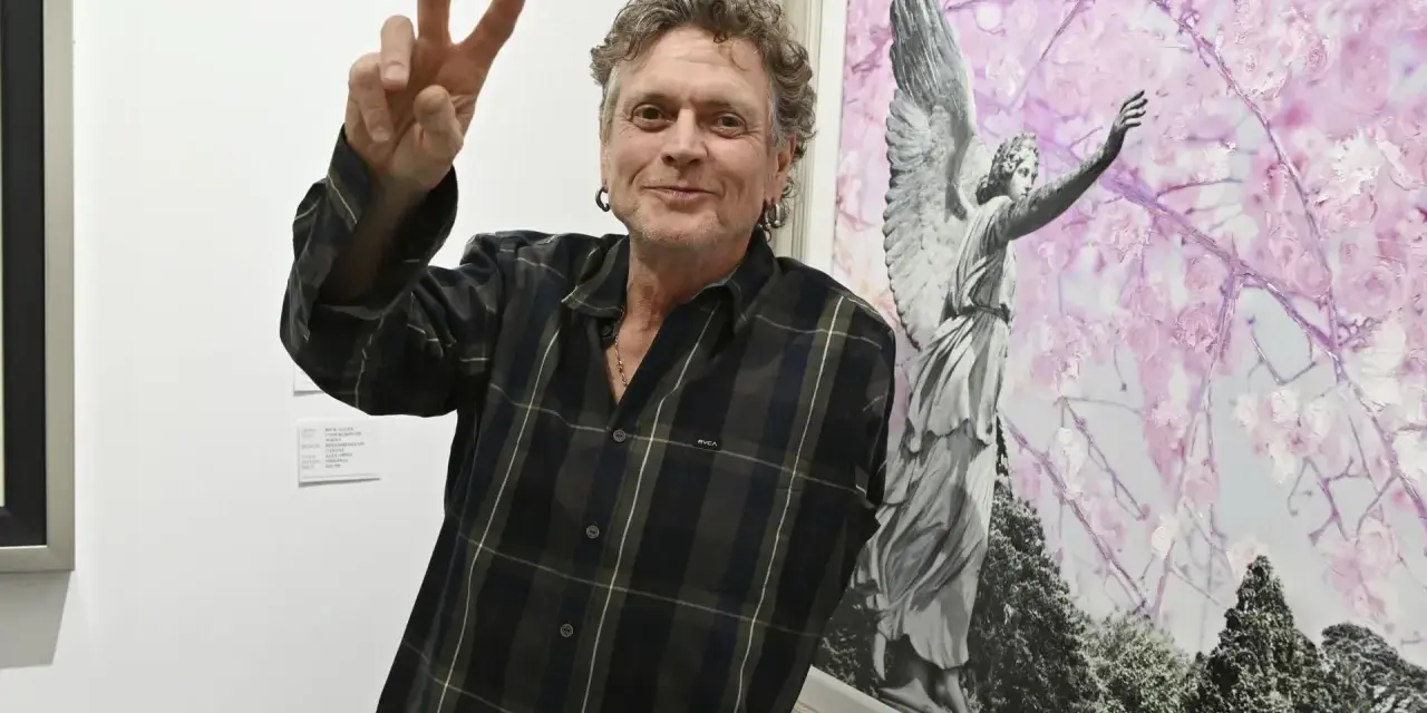 Rick Allen, perkusista Def Leppard, pozuje z uniesionym znakiem pokoju obok obrazu z aniołem i kwitnącymi drzewami.
