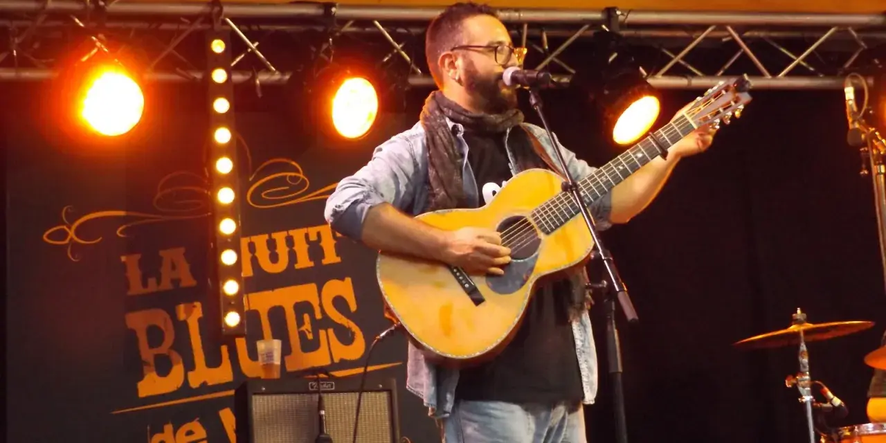 Un homme joue de la guitare sur scène, éclairé par des projecteurs. Le panneau derrière lui annonce "La Nuit du Blues", évoquant la naissance du blues.