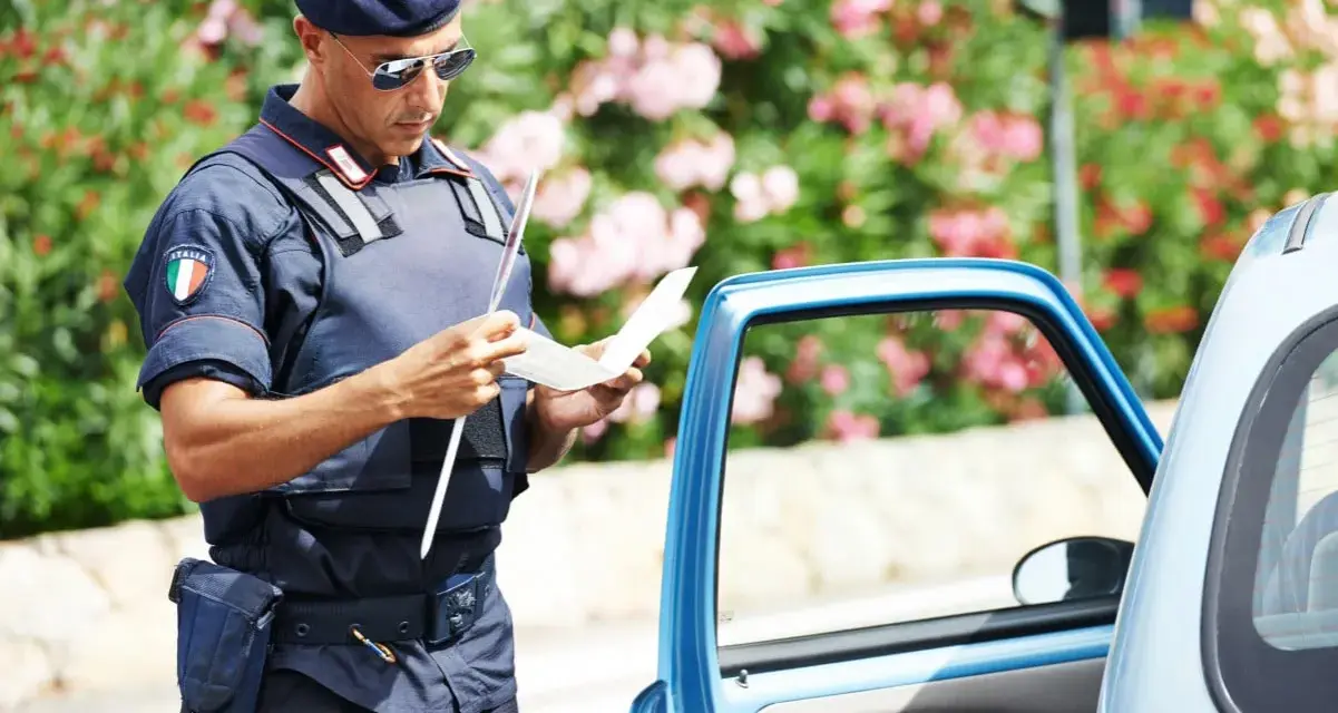 Agente di polizia italiana con giubbotto antiproiettile e berretto blu, controlla un verbale vicino a un'auto blu. Potrebbe essere un controllo legato all'art. 180 cds.