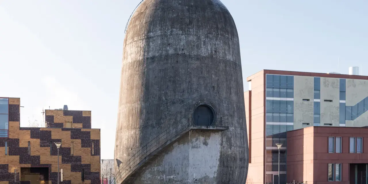 Der Trudelturm Berlin, ein futuristisches Betonbauwerk, steht neben modernen Gebäuden. Eine Außentreppe führt zu einer runden Tür.