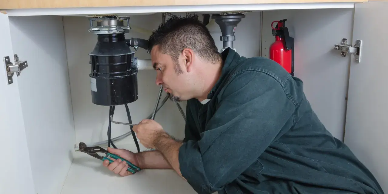 Man with pliers working on a garbage disposal under a sink.