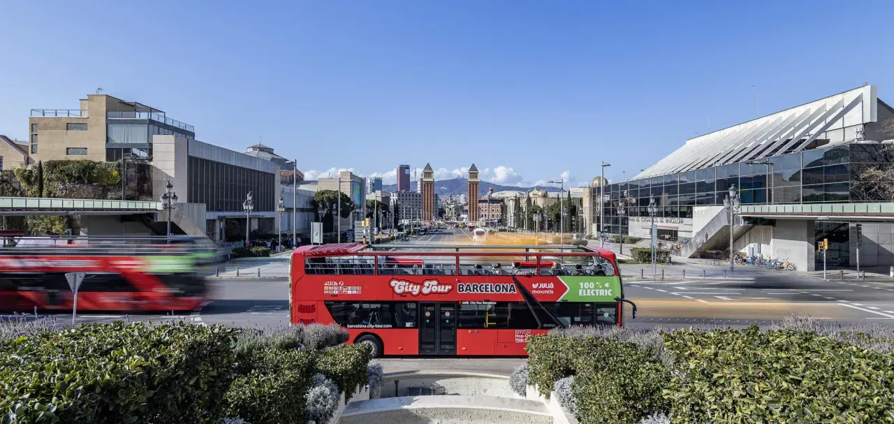 A red double-decker bus, the best hop on hop off Barcelona, drives past modern architecture under a clear blue sky.